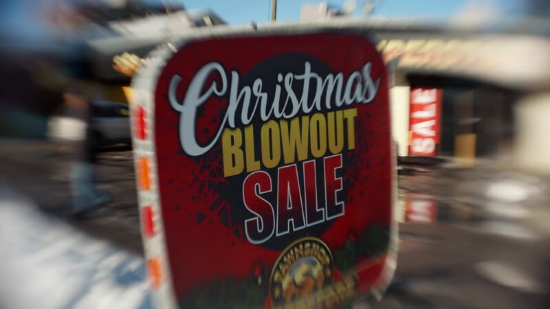 A large red sign outdoors reads "Christmas Blowout Sale" in white, yellow, and black letters. Snow is visible on the ground. The background shows another "SALE" sign and a blurred building.