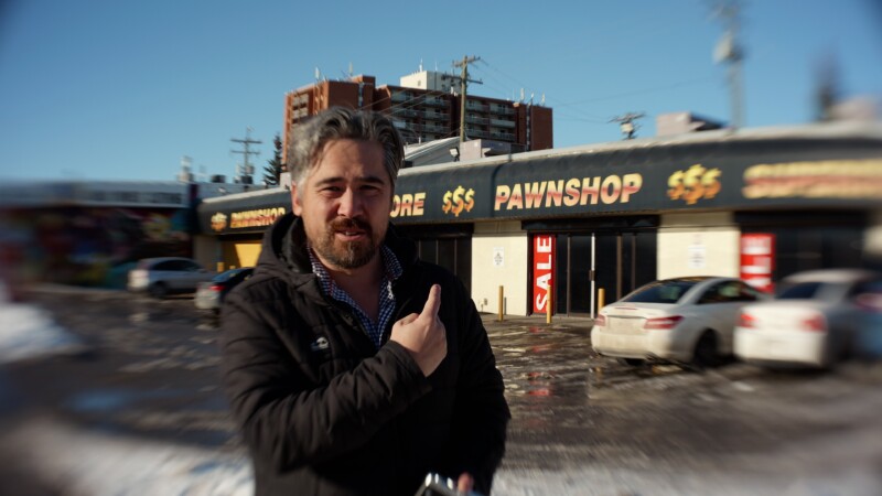 A man with gray hair and a beard stands outside in a snowy parking lot, pointing toward a pawn shop with large "PAWNSHOP $$$" letters on the storefront. Several parked cars and apartment buildings are visible in the background.