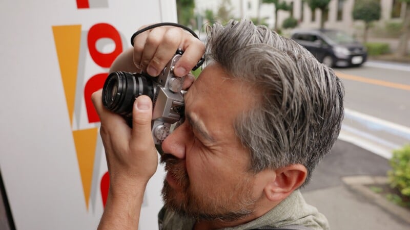 A man with gray hair and a beard holds a camera up to his eye, taking a photo outdoors near a white and red sign, with a street and parked cars visible in the background.