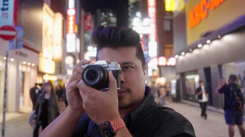 A man with dark hair holds a camera up to his face, preparing to take a photo on a brightly lit, busy city street at night with neon signs and people in the background.