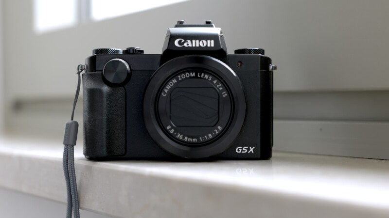 A black Canon G5 X digital camera with a zoom lens sits on a white windowsill, with a wrist strap attached to the side. The background is softly blurred.