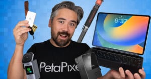 A smiling man in a "PetaPixel" shirt holds various tech gadgets, including an external drive, a camera, a battery, a tripod, and an Apple iPad with a keyboard, against a blue geometric background.