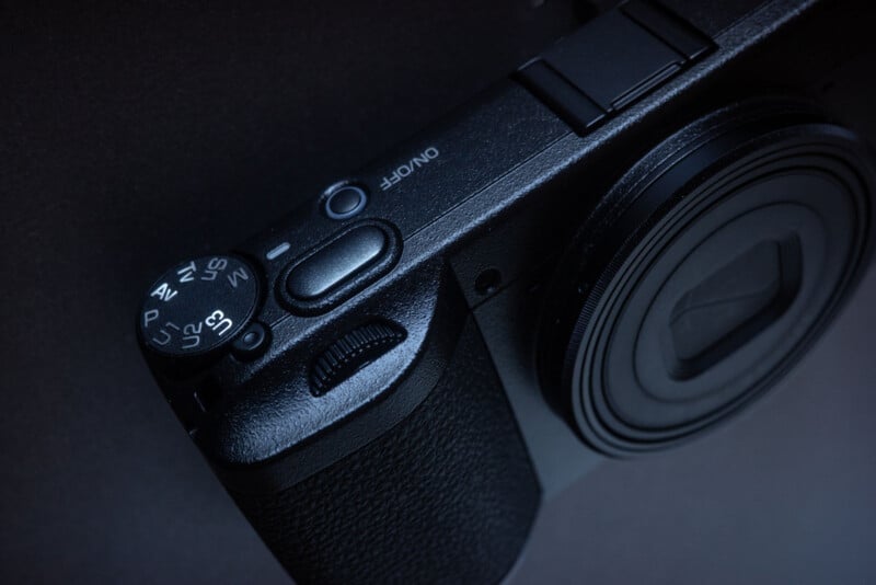 Close-up of a black digital camera showing the power button, mode dial with various settings, textured grip, and part of the lens against a dark background.