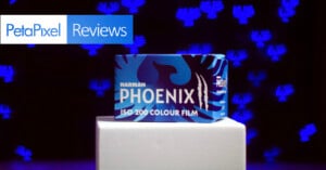 A box of Harman Phoenix ISO 200 Colour Film sits on a white platform against a dark background with blue abstract shapes, with a "PetaPixel Reviews" banner in the top left corner.