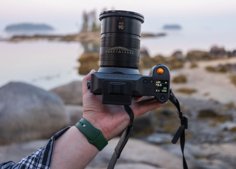 A hand holds a Hasselblad camera with a 90mm lens, displaying settings on a small screen. The background is a blurred, rocky lakeshore with water and distant trees.