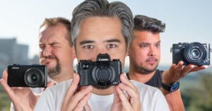 Three men hold different cameras toward the viewer. The man in the center, with gray hair, focuses on his Canon camera. The man on the left squints his eyes, holding a Sigma camera, while the man on the right holds a Sony camera.