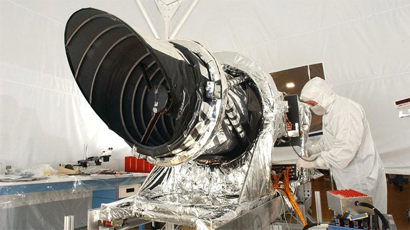 A scientist in a cleanroom suit works on a large, black and silver space telescope or instrument covered in reflective material, set up on a lab table with equipment in the background.