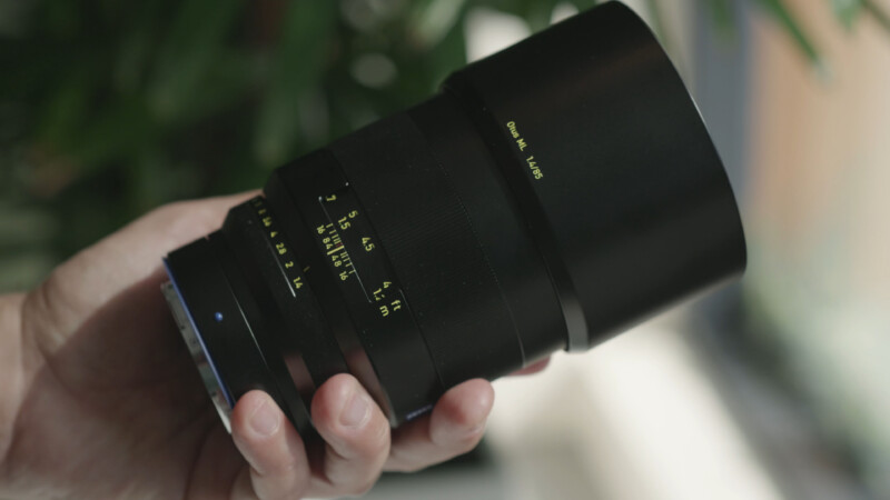 A person holds a black camera lens with yellow markings and numbers. The lens features a wide focus ring, and blurred greenery is visible in the background.