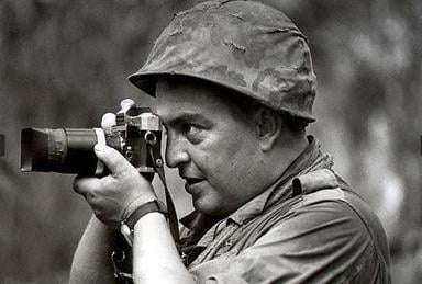 A man in military attire and a helmet holds a camera to his eye, appearing to take a photograph. The image is in black and white, with a blurred background.