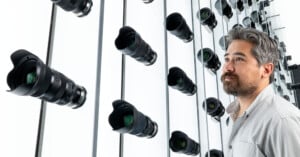 A man with gray hair and a beard stands in front of a wall lined with rows of camera lenses, some of which are reflected in the mirrored surface behind him.