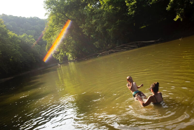Two people play in a sunlit, tree-lined river; one person is holding the other’s legs as they float on their back. A rainbow light streak shines across the scene, creating a warm, playful atmosphere.