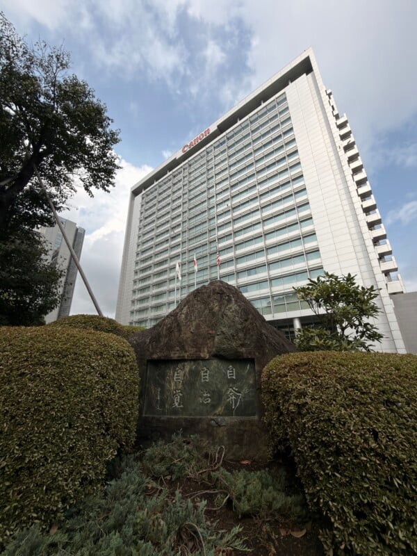 A tall, modern building with the Canon logo at the top rises behind a stone monument engraved with Japanese characters, surrounded by neatly trimmed bushes and greenery under a partly cloudy sky.
