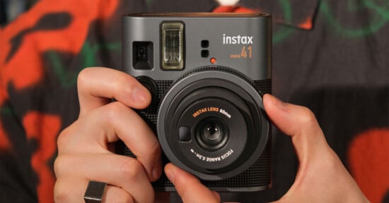 A person holds a black Instax Mini 40 instant camera, showing the lens and flash. The camera has a textured body, and the person is wearing a patterned shirt and a ring on their right hand.
