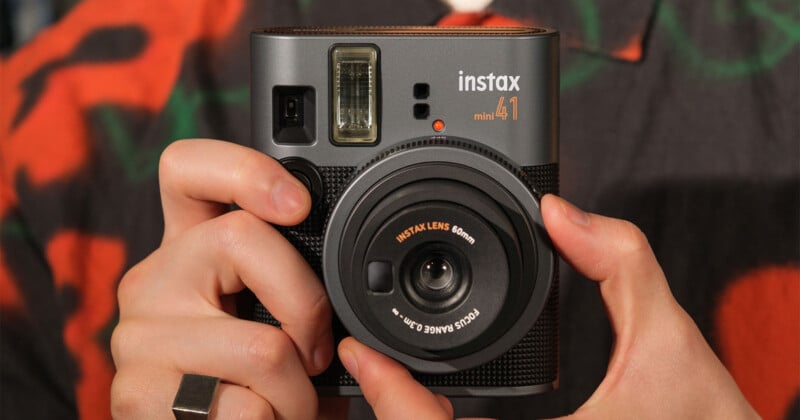 A person holds a black Instax Mini 40 instant camera, showing the lens and flash. The camera has a textured body, and the person is wearing a patterned shirt and a ring on their right hand.
