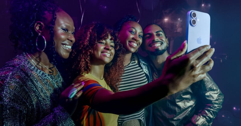 Four friends smile and pose closely together for a selfie, using a smartphone in a dimly-lit, colorful party setting with balloons in the background.