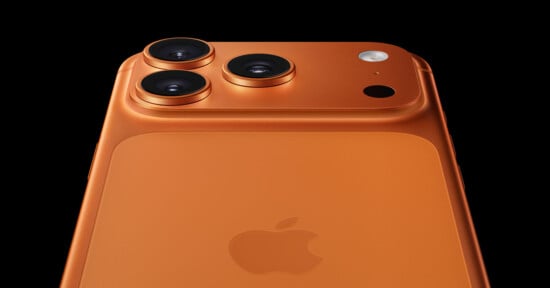 Close-up of the back of an orange iPhone, showing three camera lenses, a flash, and the Apple logo against a black background.