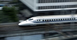 A white high-speed bullet train marked "JR 700" travels quickly along railway tracks in an urban area, with motion blur emphasizing its speed.