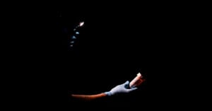 A person wearing blue gloves holds a prescription pill bottle in one hand and examines it with a flashlight in the other, set against a completely dark background.