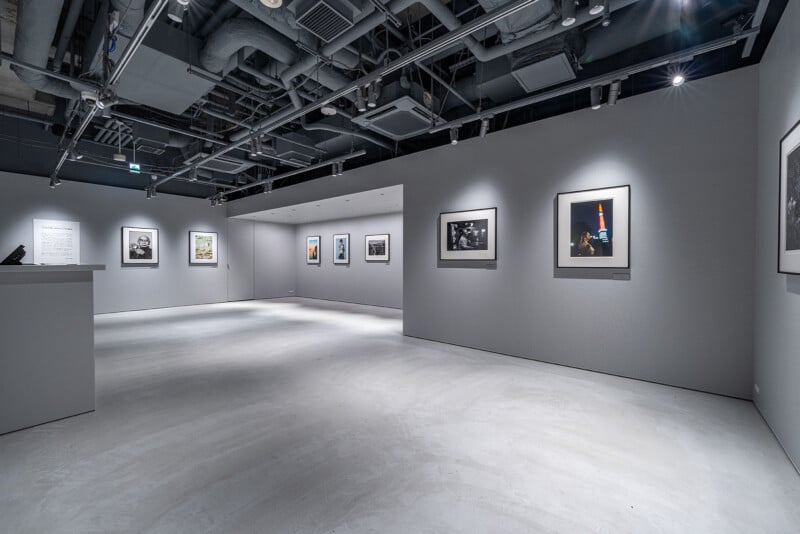 A modern art gallery with gray walls and ceiling, featuring framed photographs displayed on the walls under focused spotlights. The gallery has a minimalist, industrial design with exposed pipes.