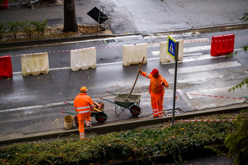 Two workers in orange safety clothing work on a city street near a crosswalk. One is mixing concrete in a wheelbarrow, while the other stands nearby. Red and white barriers and tape mark off the area.