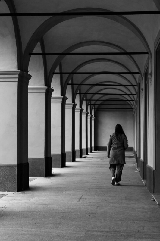 A person walks alone under a series of tall, arched columns in a covered walkway. The scene is in black and white, creating a calm, contemplative atmosphere with strong shadows and architectural lines.