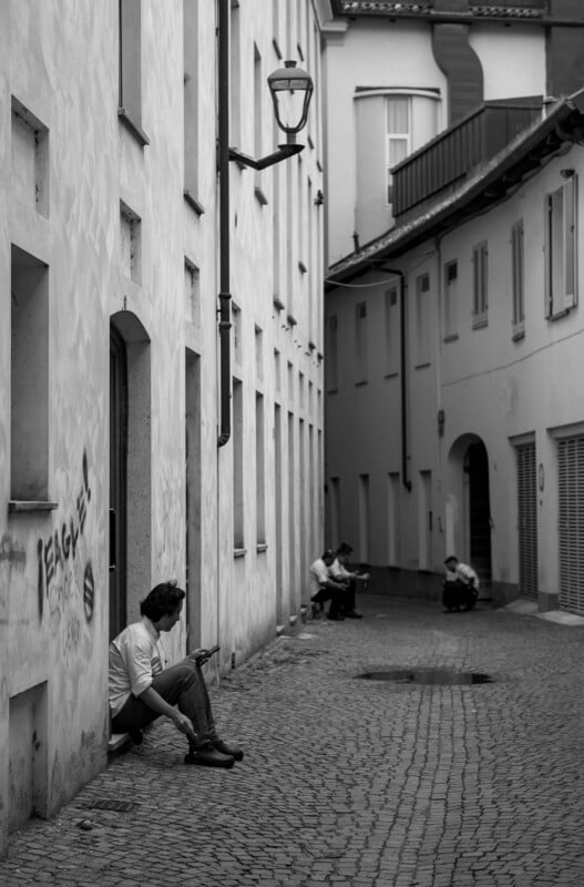 A narrow cobblestone alleyway with several people sitting against the white walls of buildings, some looking at their phones. The scene is in black and white, with graffiti visible on one wall.