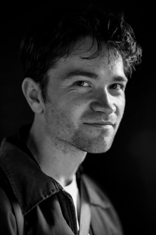 Black and white portrait of a young man with short, dark hair, slight stubble, and a subtle smile, wearing a collared jacket and looking slightly to the side against a dark background.