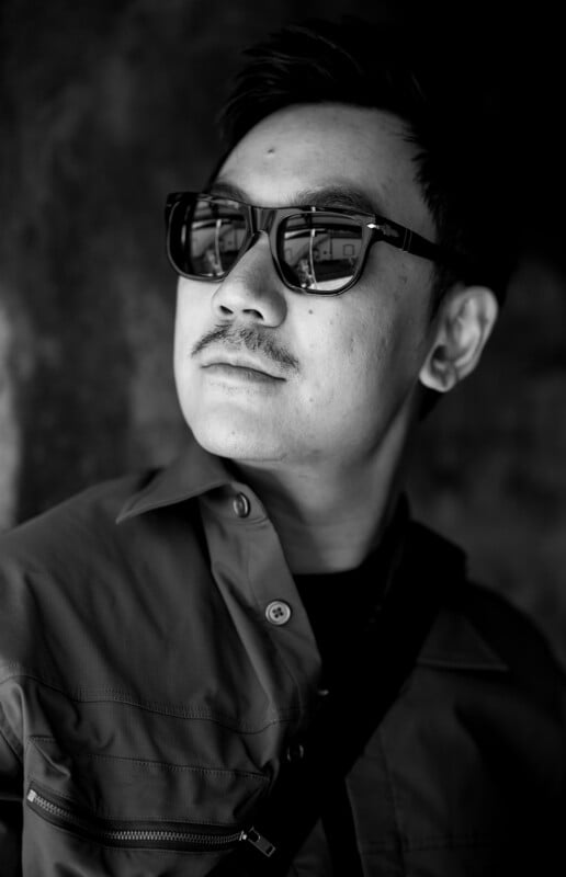 A man with a mustache wearing sunglasses and a button-up jacket looks to the side. The black-and-white photo has a shadowy background and highlights reflections in his sunglasses.