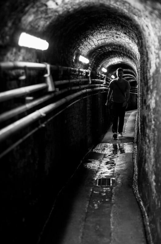A person walks alone through a narrow, dimly lit underground tunnel with exposed pipes along the wall and water on the floor. The scene is in black and white.