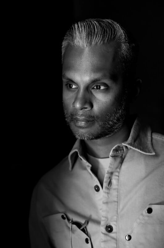 Black and white portrait of a man with short, neatly styled gray hair and a trimmed beard, wearing a button-up shirt and looking thoughtfully to the side. The background is dark, highlighting his face.