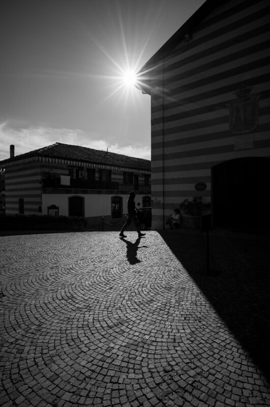 A person walks across a cobblestone square, casting a long shadow in the sunlight. The sun is shining brightly above a striped building, creating strong contrasts of light and shadow.