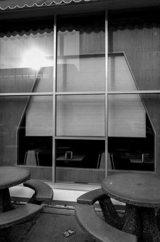 Black and white photo of an empty diner seen through a large window with unique geometric blinds. In the foreground are round concrete picnic tables. Sunlight reflects off the glass, and the interior appears deserted.