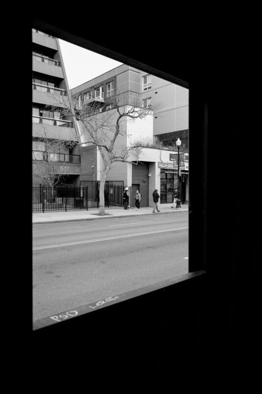 A city street view is framed by a dark window. Four people walk on the sidewalk beside buildings and leafless trees. The photo is in black and white.