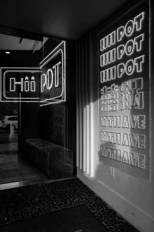 Black and white photo of a neon sign reading “Hii Pot” and “17th Ave” in English and Chinese characters, illuminating a modern entryway with a reflection in a glass wall and a glimpse of the street outside.