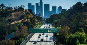 Cars drive along a freeway lined with green hills toward downtown Los Angeles, with tall skyscrapers visible in the background and green highway signs overhead directing traffic.