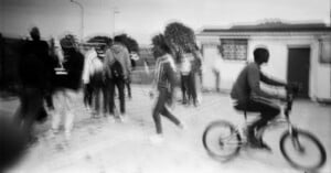A black-and-white, motion-blurred photo shows a group of people walking on the left and a person riding a bicycle on the right, outside near buildings. The scene feels dynamic and slightly unfocused.