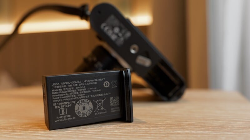 A close-up of a Leica rechargeable lithium-ion battery standing upright on a wooden surface, with a partially blurred camera and its battery compartment in the background.