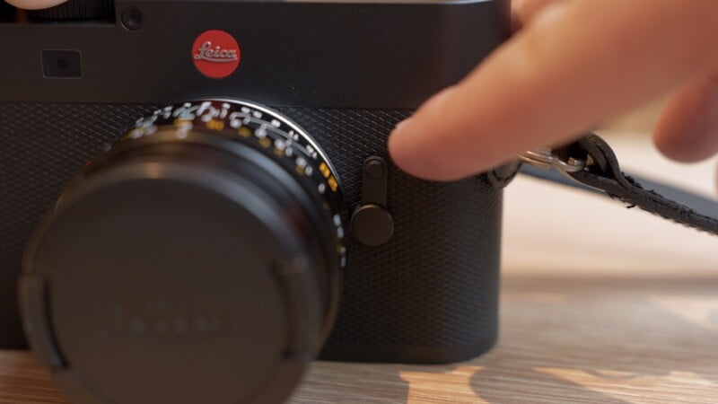 A close-up of a person’s hand adjusting a dial on a black Leica camera with a lens cap on, placed on a wooden surface.