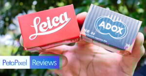 A hand holds two boxes of camera film: a red Leica box and a blue-striped Adox HR 50 box. The image features a PetaPixel Reviews banner in the lower left corner, with greenery in the blurred background.