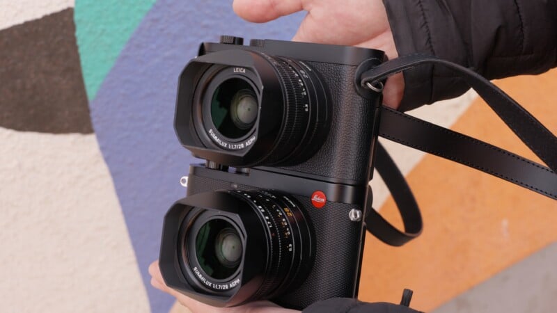 Close-up of hands holding a unique double-lens Leica camera in front of a colorful, abstract painted wall. The camera features two large lenses stacked vertically, and a black strap is visible.