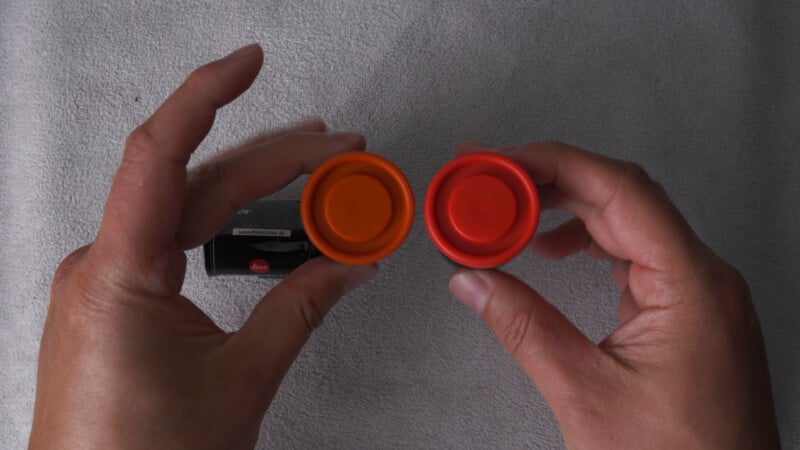 Two hands holding orange and red circular caps above a gray surface, with a black rectangular object partially visible on the left side.