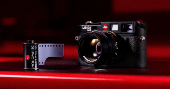 A Leica M6 film camera sits on a red surface next to a roll of Leica Monochrom 400 black and white 35mm film, with a dark, moody background.