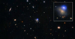 A deep space image showing numerous galaxies and stars scattered across a dark background, with an inset zoom highlighting a bright blue glowing area within a specific galaxy.