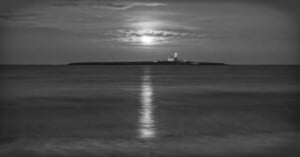 A black and white photo of a small island with a lighthouse, illuminated by the moonlight. The moon is partially covered by clouds, and its reflection shimmers on the calm ocean water in the foreground.