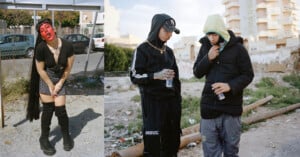 A person with red face paint, black outfit, and boots poses near a fence. Nearby, two people in hooded jackets and streetwear stand on rough ground, drinking water, with buildings in the background.