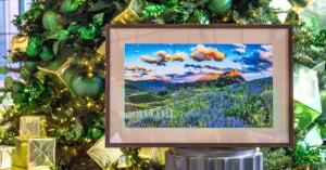 A framed landscape photo of a colorful mountain field with wildflowers is displayed in front of a decorated Christmas tree with green ornaments and gold gift boxes.