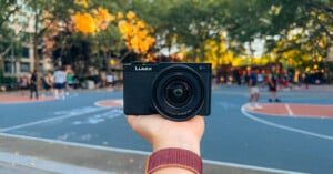 A hand holds a black Lumix camera in focus, with an outdoor basketball court and blurred players and trees in the background.