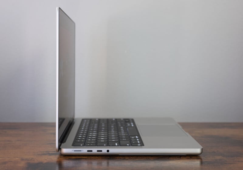 A slim, silver laptop is open to a side profile angle on a wooden surface, with its screen tilted slightly back and various ports visible along the left edge. The background is plain and light gray.