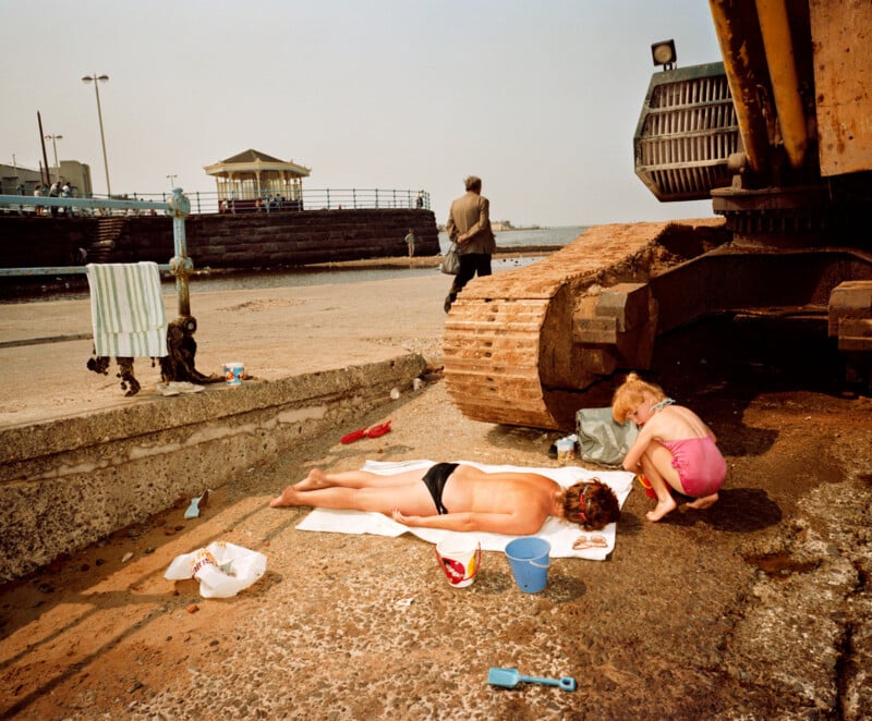 A person in swimwear lies on a towel sunbathing near a large construction vehicle; a child plays beside them with a bucket and spade. The scene is set on a concrete pier by the sea, with scattered beach toys and distant figures.