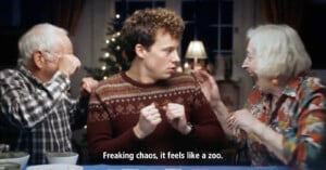 A young man in a patterned sweater sits between two lively older adults at a table, looking startled as they gesture energetically. Subtitles read, "Freaking chaos, it feels like a zoo.